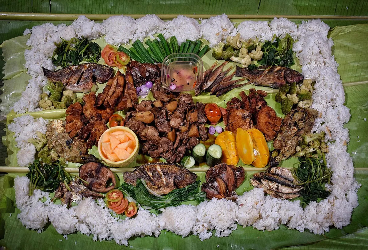 Boodle fight feast of rice, grilled seafood, pork and fruit laid out on banana leaves on a long table